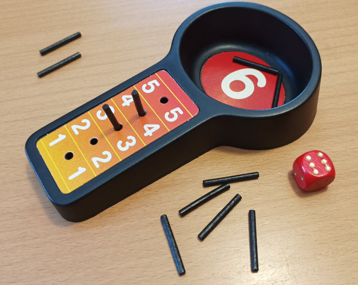 A board with five sockets, numbered 1 to 5 and a bigger hole numbered 6. In holes 3 and 4 there are short pegs. More pegs are next to the board, together with a 6-sided die.